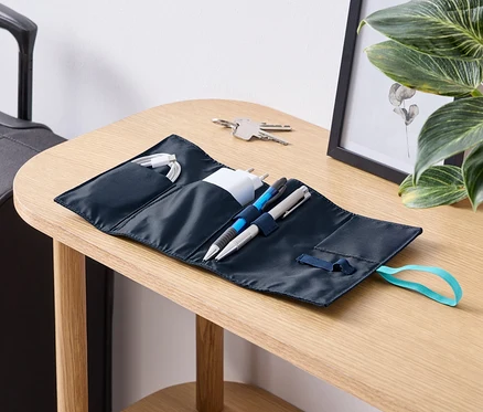 Sur une table, un organisateur de voyage pour câbles ouvert avec des câbles, un chargeur et des stylos.