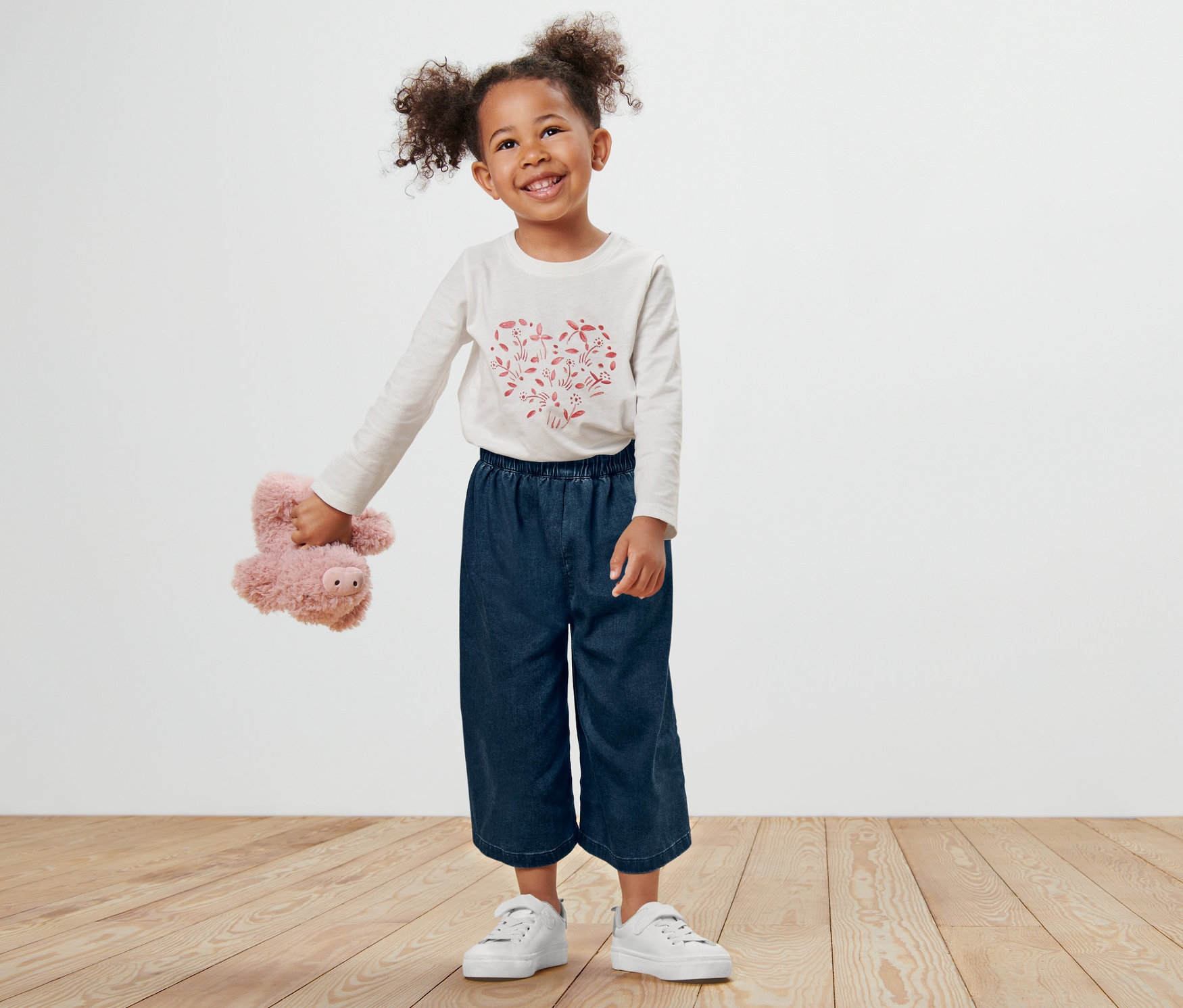 Une petite fille souriante se tient debout et tient un paresseux en peluche. Elle porte une jupe-culotte pour tout-petit, des baskets blanches et une chemise à manches longues.