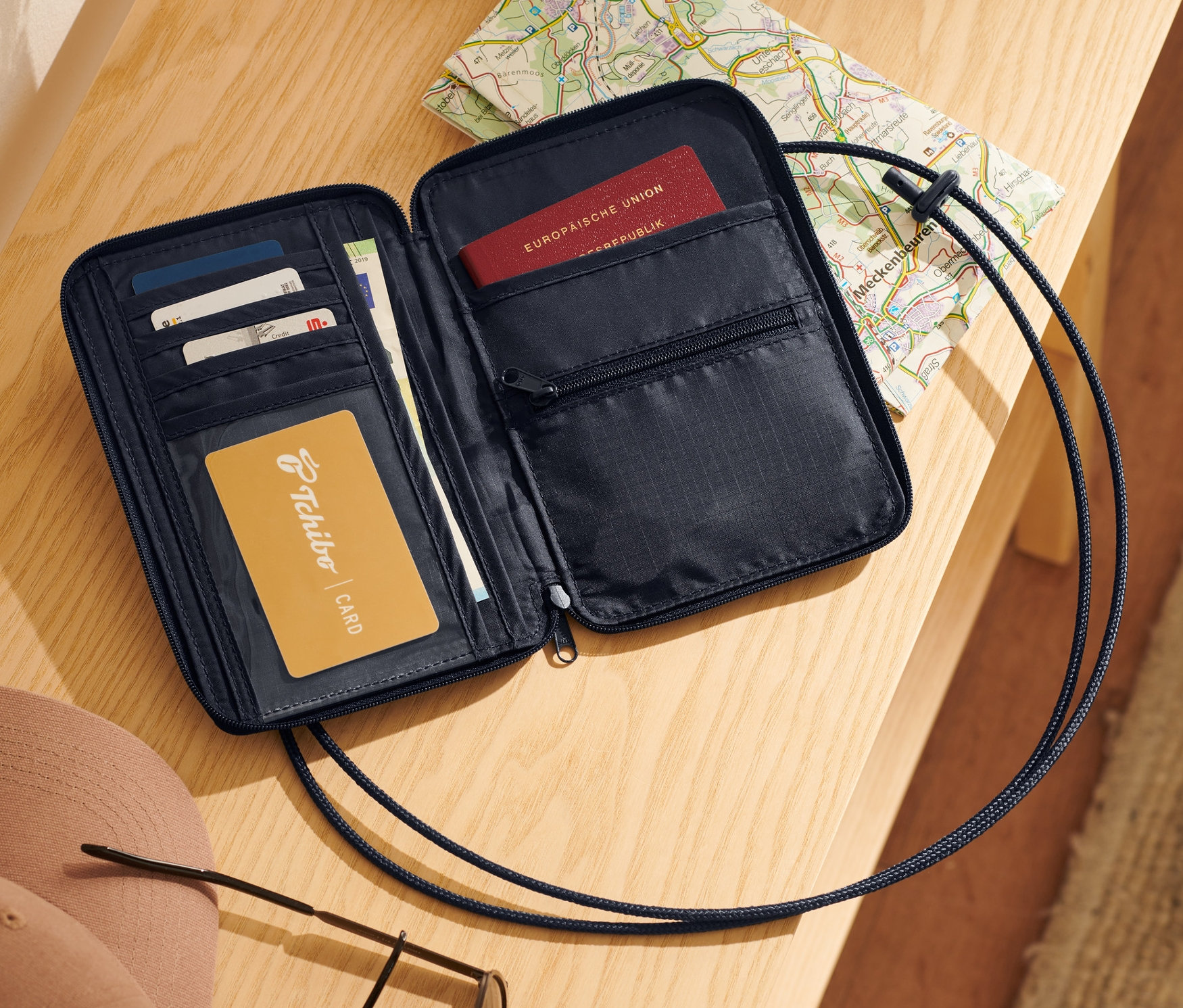 Pochette tour de cou noire ouverte avec cartes, passeport et carte sur une table en bois.