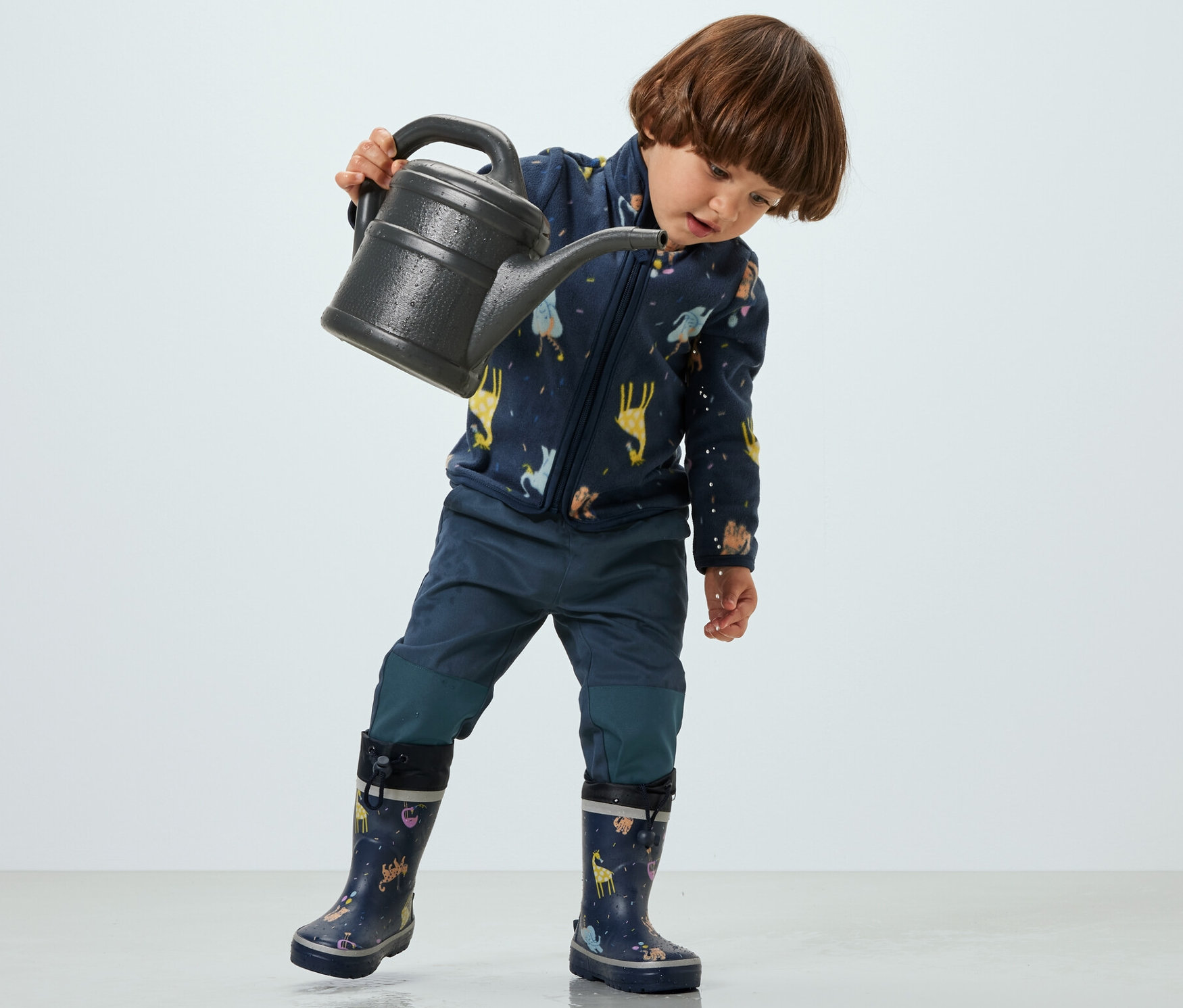 Un enfant tient un arrosoir et porte un pantalon d’extérieur pour enfant.