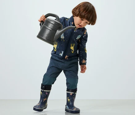 Un enfant tient un arrosoir et porte un pantalon d’extérieur pour enfant.