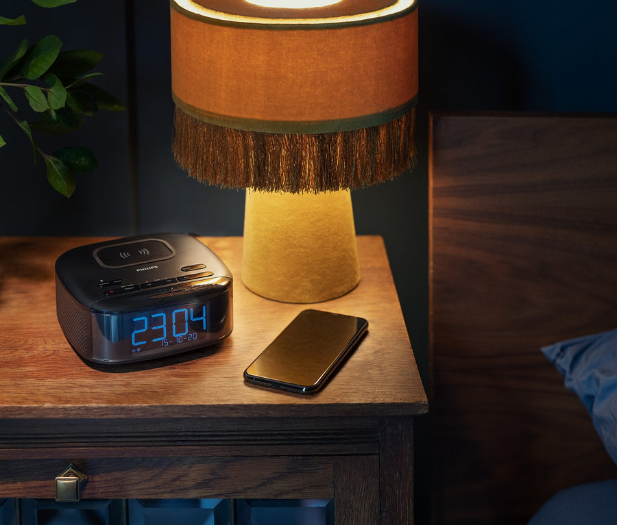 Sur une table de chevet se trouvent un radio-réveil DAB Philips « TAR7705 », un téléphone portable et une lampe.