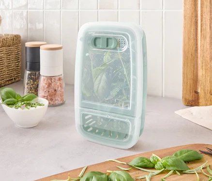 Un récipient de conservation d'herbes avec des herbes à l'intérieur est posé sur un comptoir à côté d'ingrédients et d'une planche à découper.