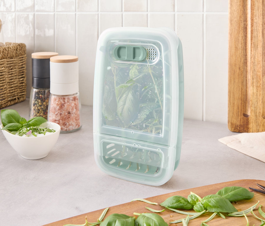 Un récipient de conservation d'herbes avec des herbes à l'intérieur est posé sur un comptoir à côté d'ingrédients et d'une planche à découper.