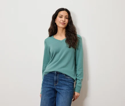 Une femme aux longs cheveux bruns pose devant un fond blanc. Elle porte un jean bleu et un pull vert.