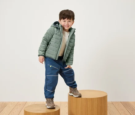 Un garçon souriant pose sur un socle en bois, vêtu d'une veste verte, d'un pantalon bleu et de chaussures marron.