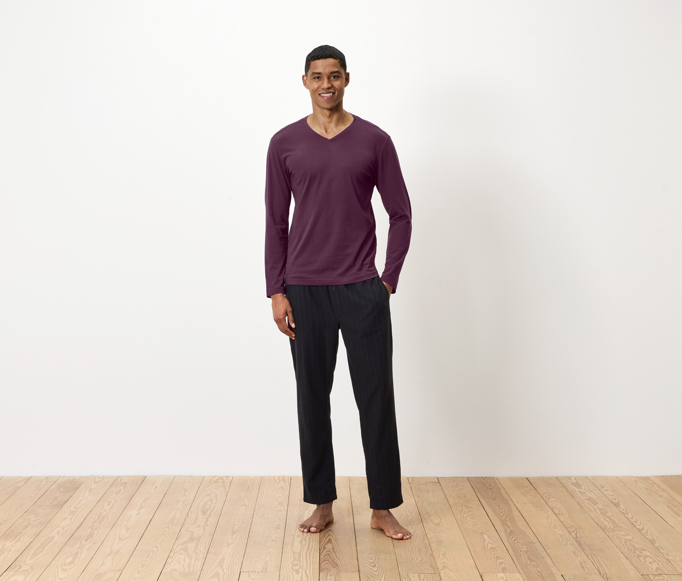 Un homme se tient pieds nus sur un plancher en bois, portant un t-shirt à manches longues bordeaux et un pantalon noir.