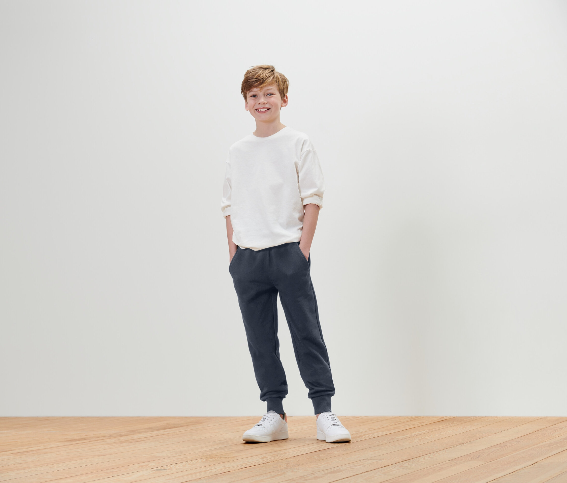 Un garçon se tient debout, les mains dans les poches, portant un t-shirt blanc, un jogging bleu et des baskets blanches.