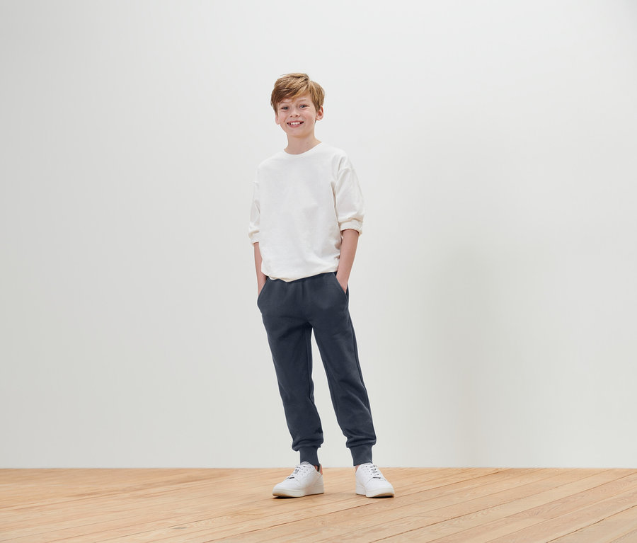 Un garçon se tient debout, les mains dans les poches, portant un t-shirt blanc, un jogging bleu et des baskets blanches.