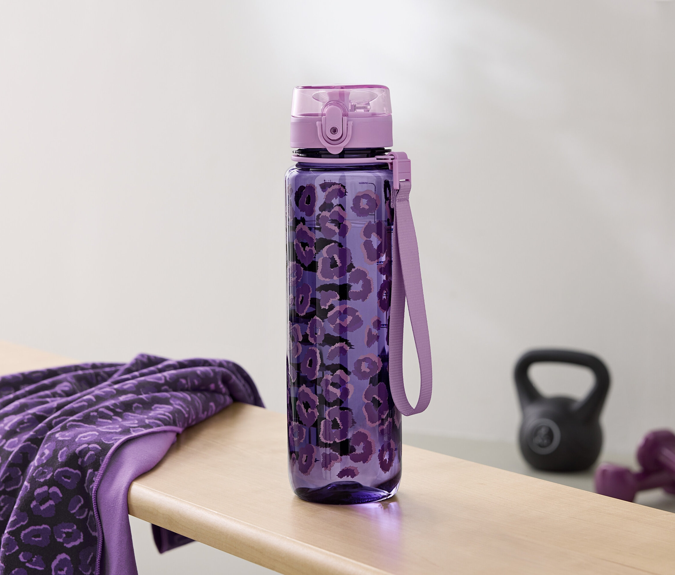 Une bouteille d'eau violette à motifs repose à côté d'une serviette et d'haltères sur un banc.