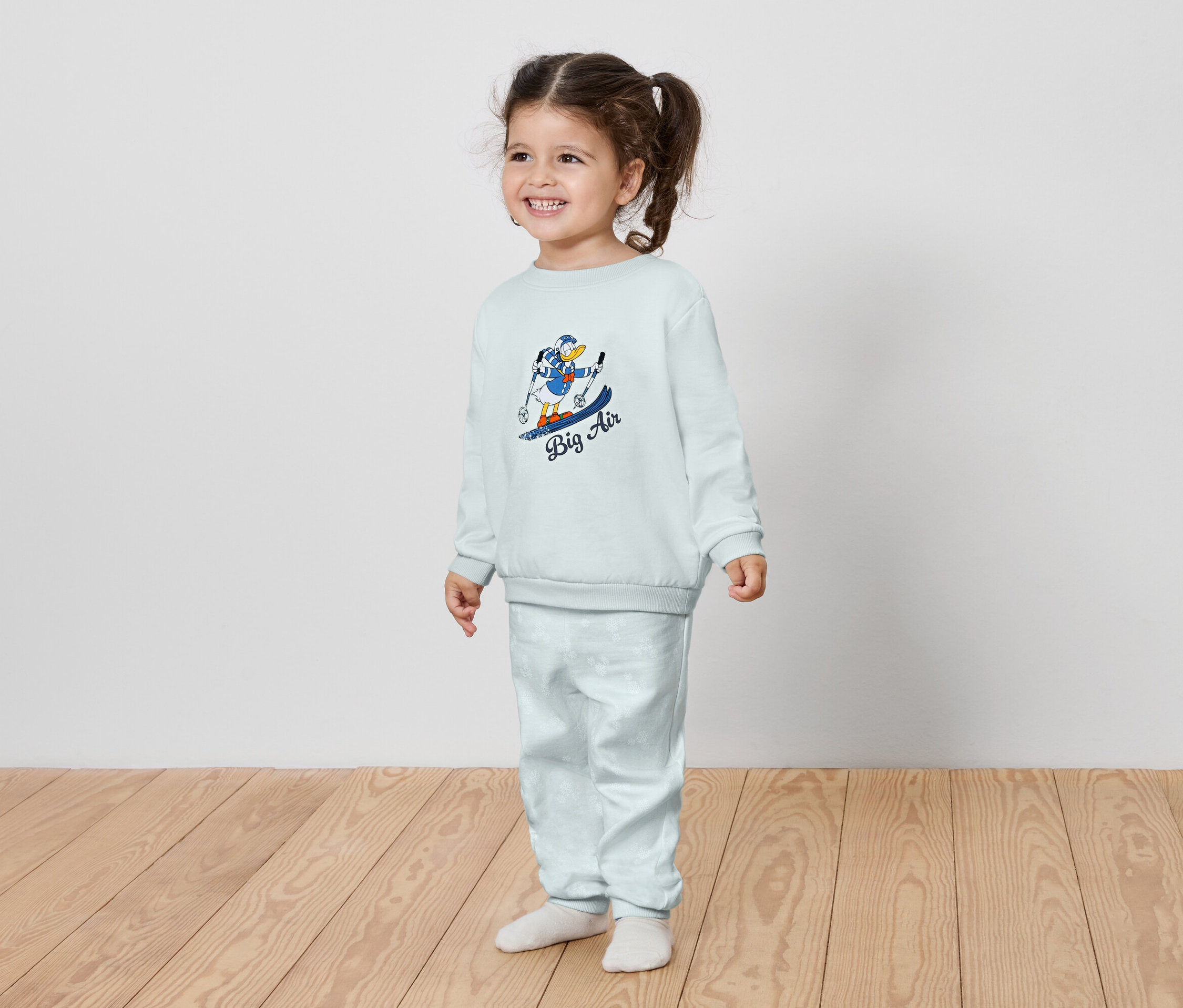 Petite fille souriante dans un ensemble bleu clair avec un imprimé de personnage de dessin animé sur un snowboard.