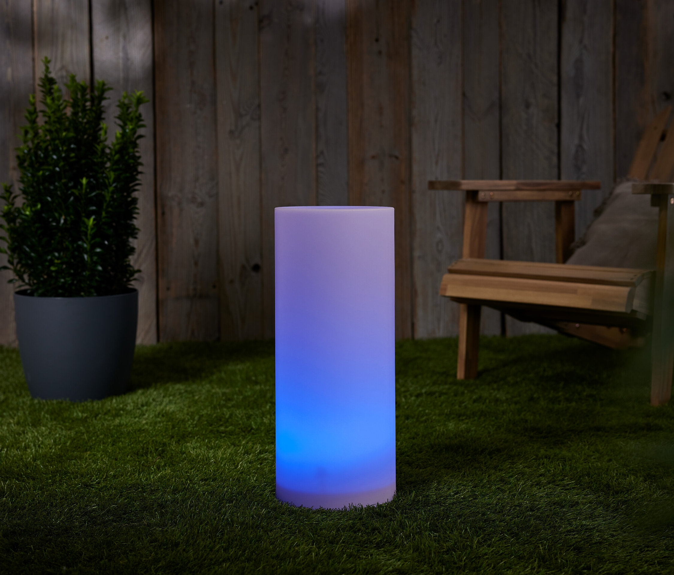 Une colonne lumineuse solaire avec changement de couleur, env. 50 cm, est posée sur l'herbe à côté d'un pot de fleurs et d'une chaise en bois.