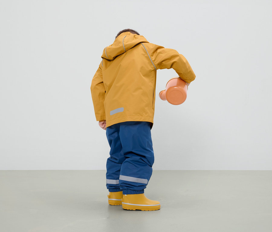 Un garçon vêtu d'une veste moutarde, d'un pantalon bleu et de bottes de pluie jaunes tient un arrosoir orange.