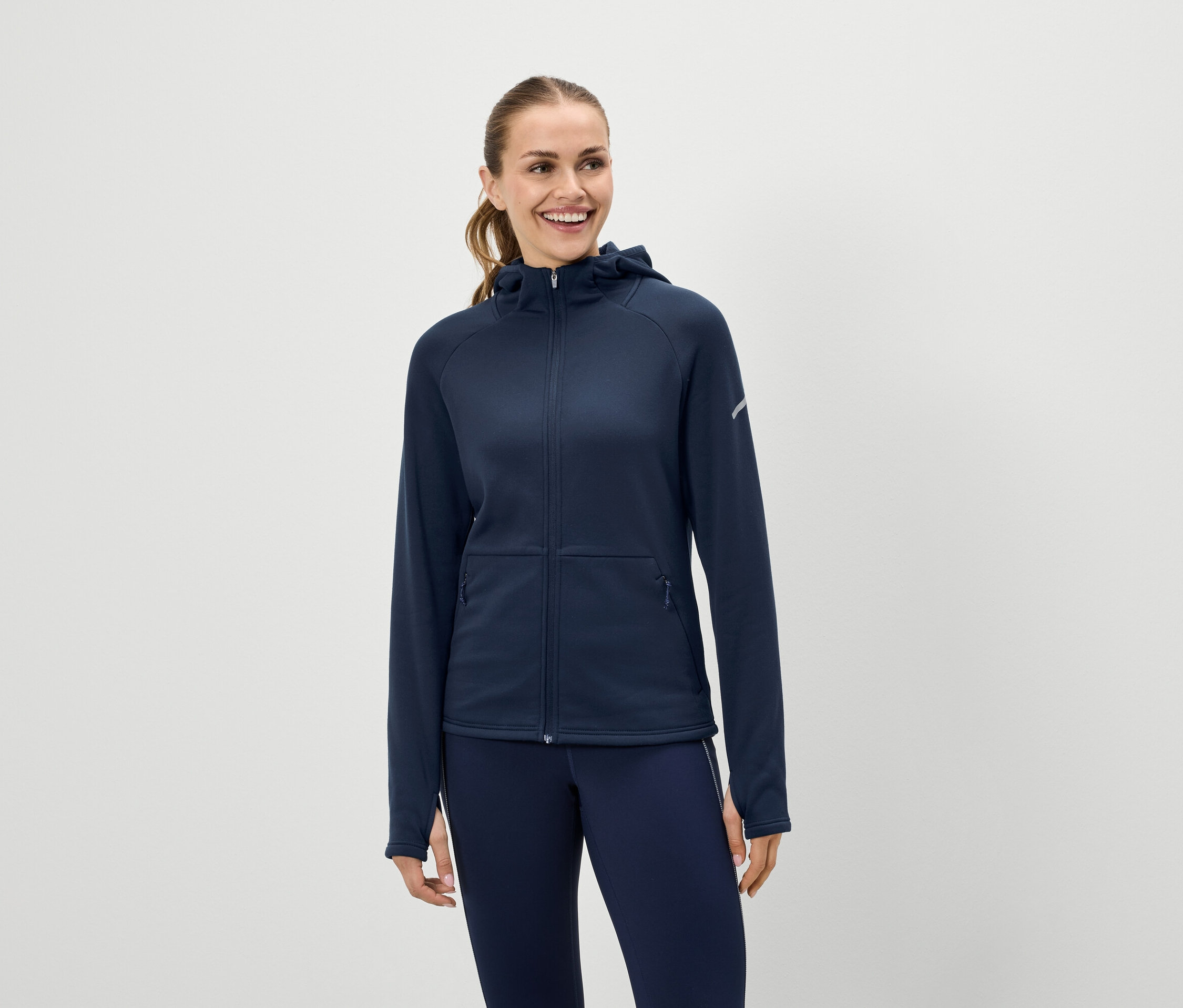 Une femme souriante porte un sweat à capuche bleu marine zippé et un legging bleu marine.