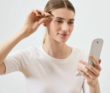 Une femme s'épile les sourcils avec une pince à épiler en regardant dans un miroir.
