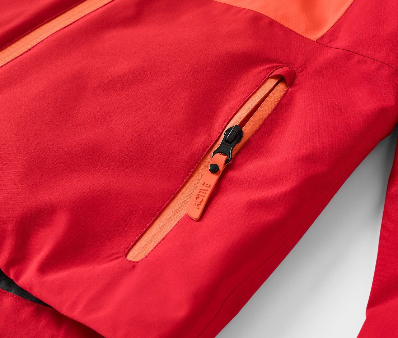Détail d'une veste rouge avec fermeture éclair orange et poche poitrine.