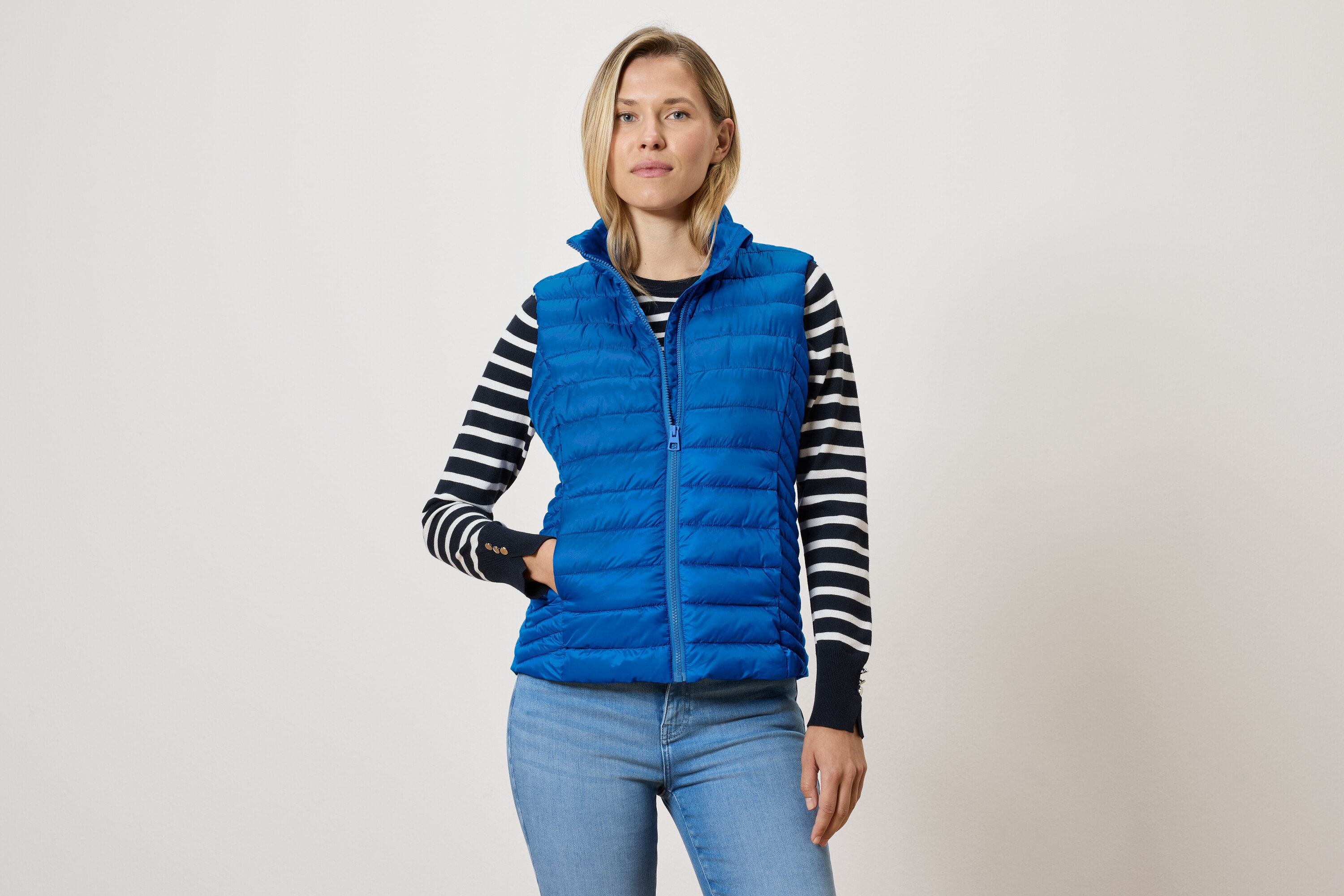 Une femme portant une veste sans manches matelassée bleue avec fermeture éclair, un pull rayé noir et blanc et un jean bleu.