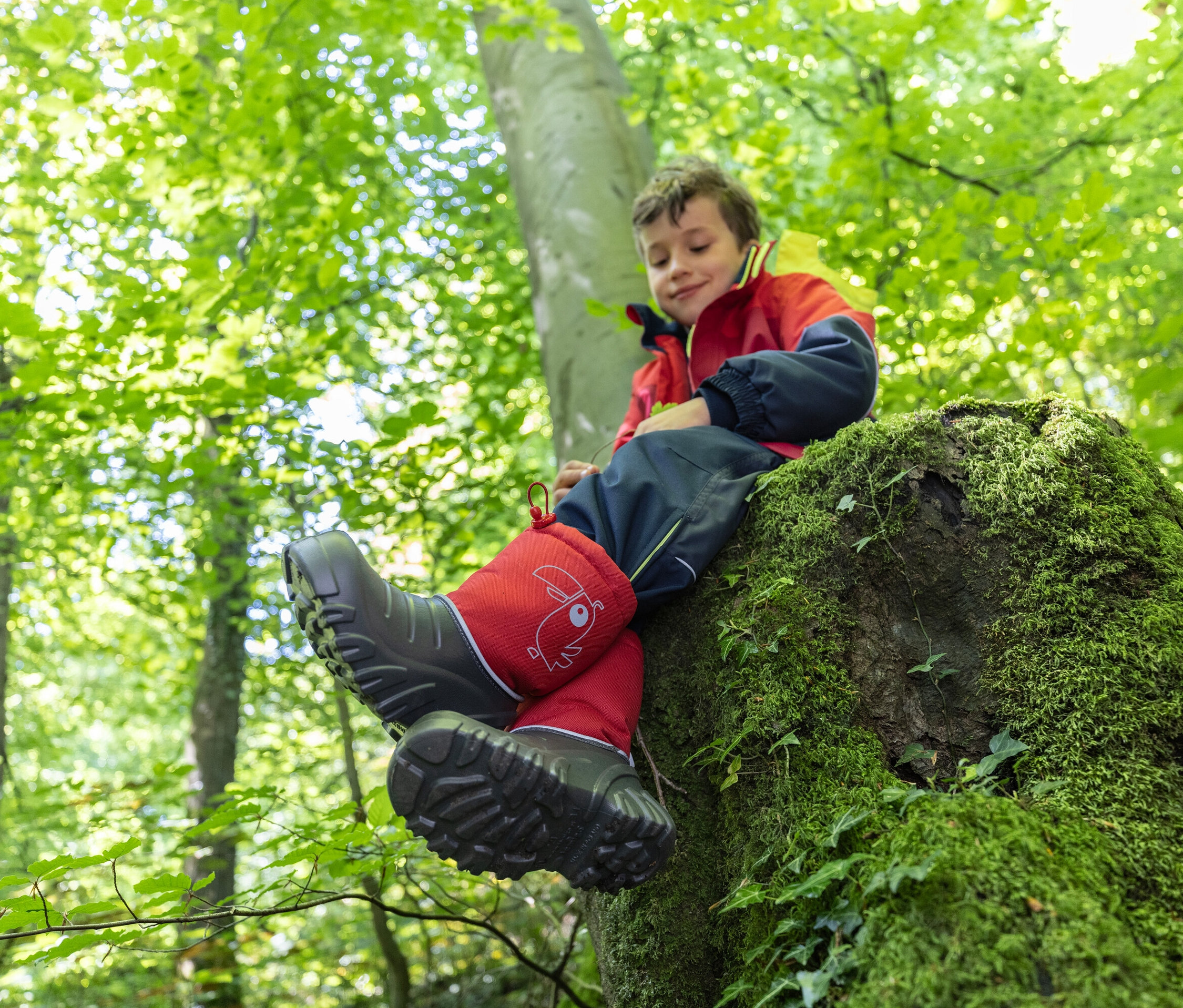 Un garçon est assis sur une souche d'arbre couverte de mousse dans une forêt et porte des bottes en caoutchouc rouges.