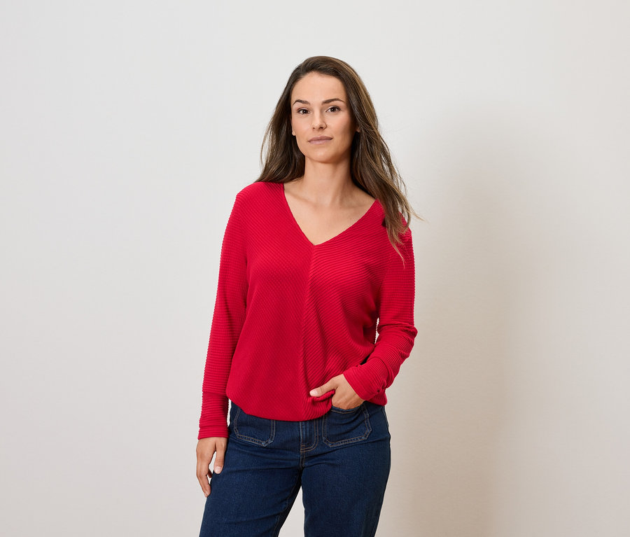 Femme aux cheveux bruns posant en pull rouge à manches longues et jean bleu.