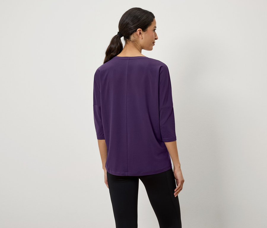 Une femme aux cheveux foncés attachés en queue de cheval, vue de dos, porte un t-shirt violet à manches trois-quarts et un legging noir.