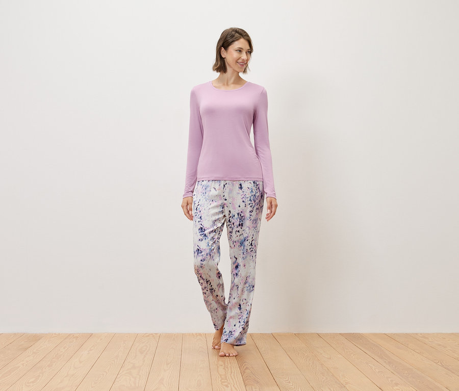 Une femme se tient pieds nus sur un plancher en bois, portant un pull violet et un pantalon imprimé.