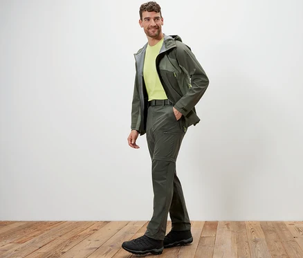 Un homme pose en pantalon technique 2 en 1 kaki et veste sur un plancher en bois.