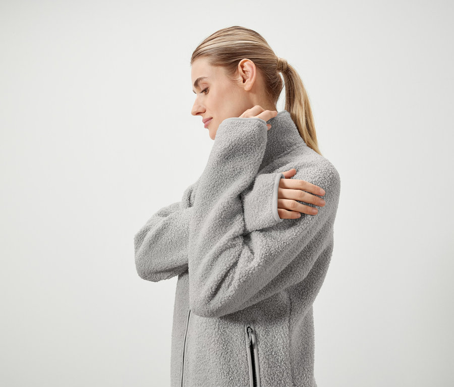 Profil d'une femme blonde aux cheveux attachés en queue de cheval portant une veste grise duveteuse à fermeture éclair sur fond blanc.
