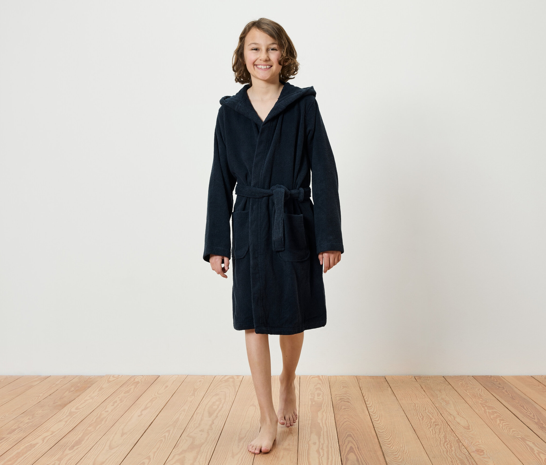 Une adolescente souriante marche pieds nus sur un plancher en bois, portant un peignoir bleu marine à capuche.