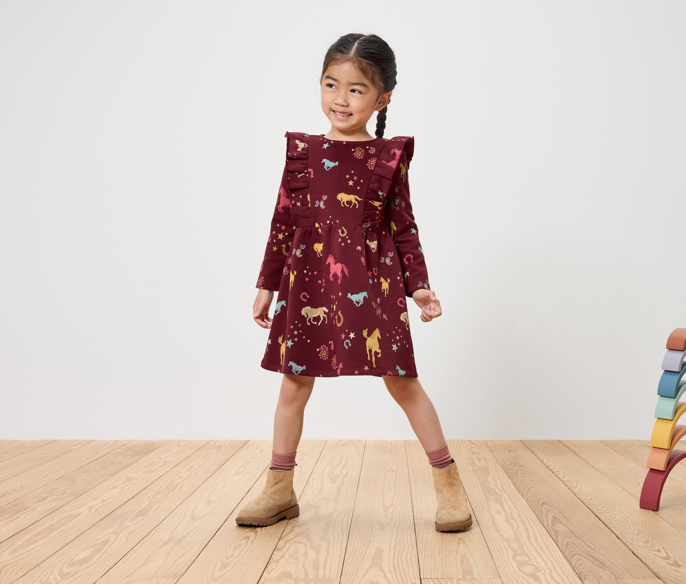 Fillette souriante vêtue d'une robe bordeaux à imprimé cheval et de bottes marron se tenant sur un plancher en bois à côté d'un jouet en bois arc-en-ciel.