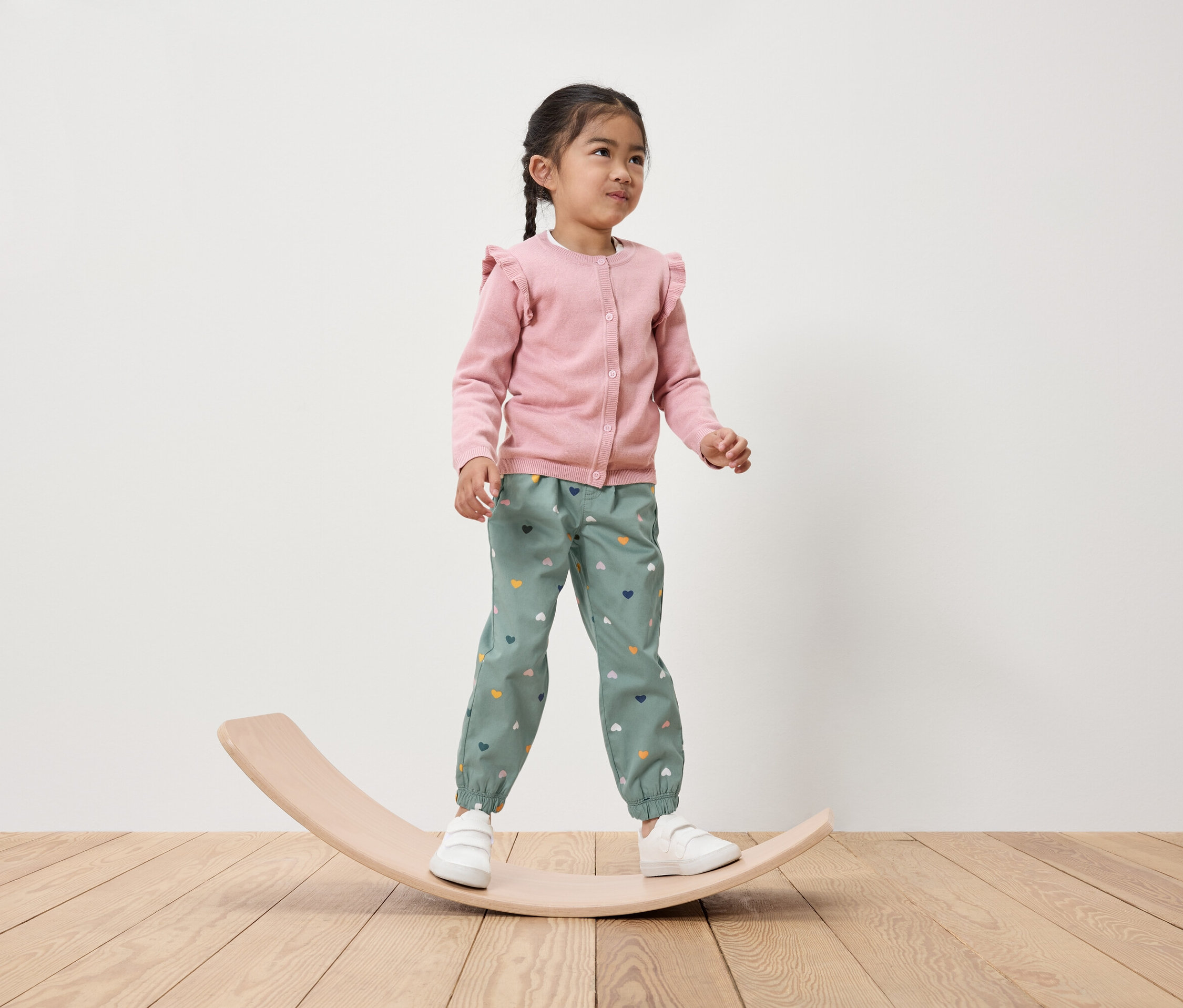 Une fillette se tient sur une planche d'équilibre en bois, portant un cardigan rose et un pantalon vert avec des cœurs.