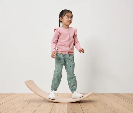 Une fillette se tient sur une planche d'équilibre en bois, portant un cardigan rose et un pantalon vert avec des cœurs.