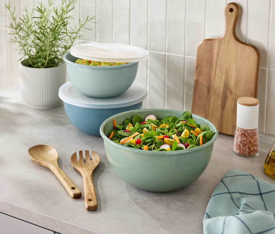 Sur un comptoir de cuisine se trouvent un saladier, des couverts en bois, une salière, une bouteille d'huile, une planche à découper et une plante en pot.