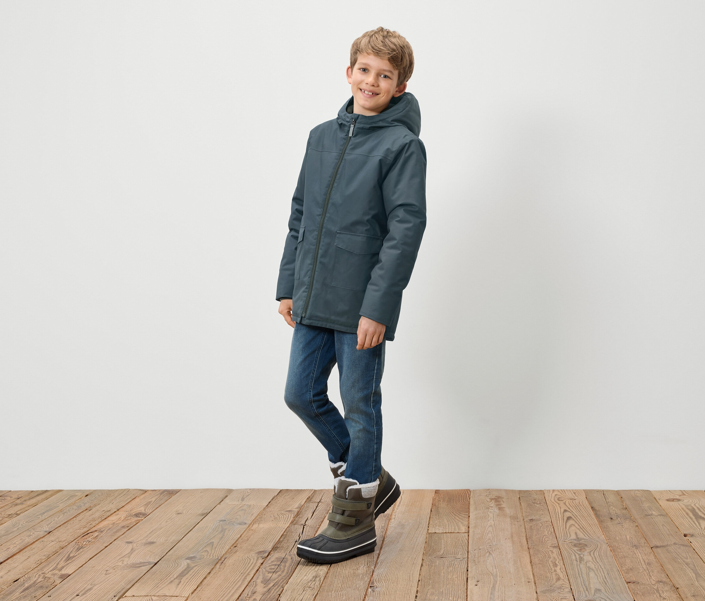 Garçon posant en veste à capuche bleue, jean et bottes d'hiver.