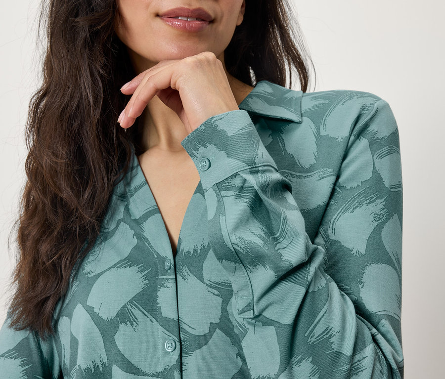 Haut du corps d'une femme brune portant un chemisier vert à motifs, la main au menton.
