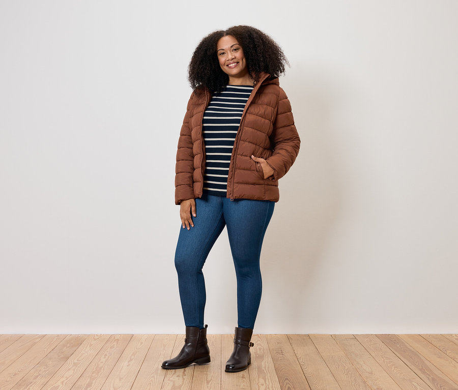 Une femme aux cheveux bouclés pose en doudoune marron, jean bleu et bottes marron.