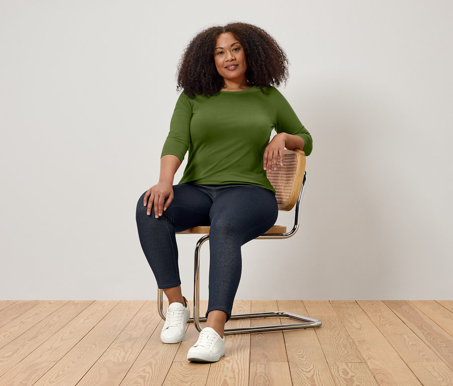 Une femme aux cheveux bouclés est assise sur une chaise, portant un t-shirt vert à manches trois-quarts, un jean foncé et des baskets blanches.