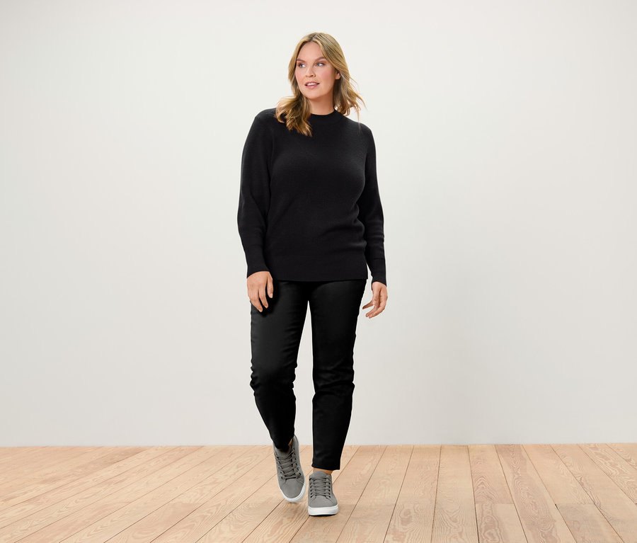 Une femme marchant sur un plancher en bois porte un pull noir, un pantalon noir et des baskets grises.