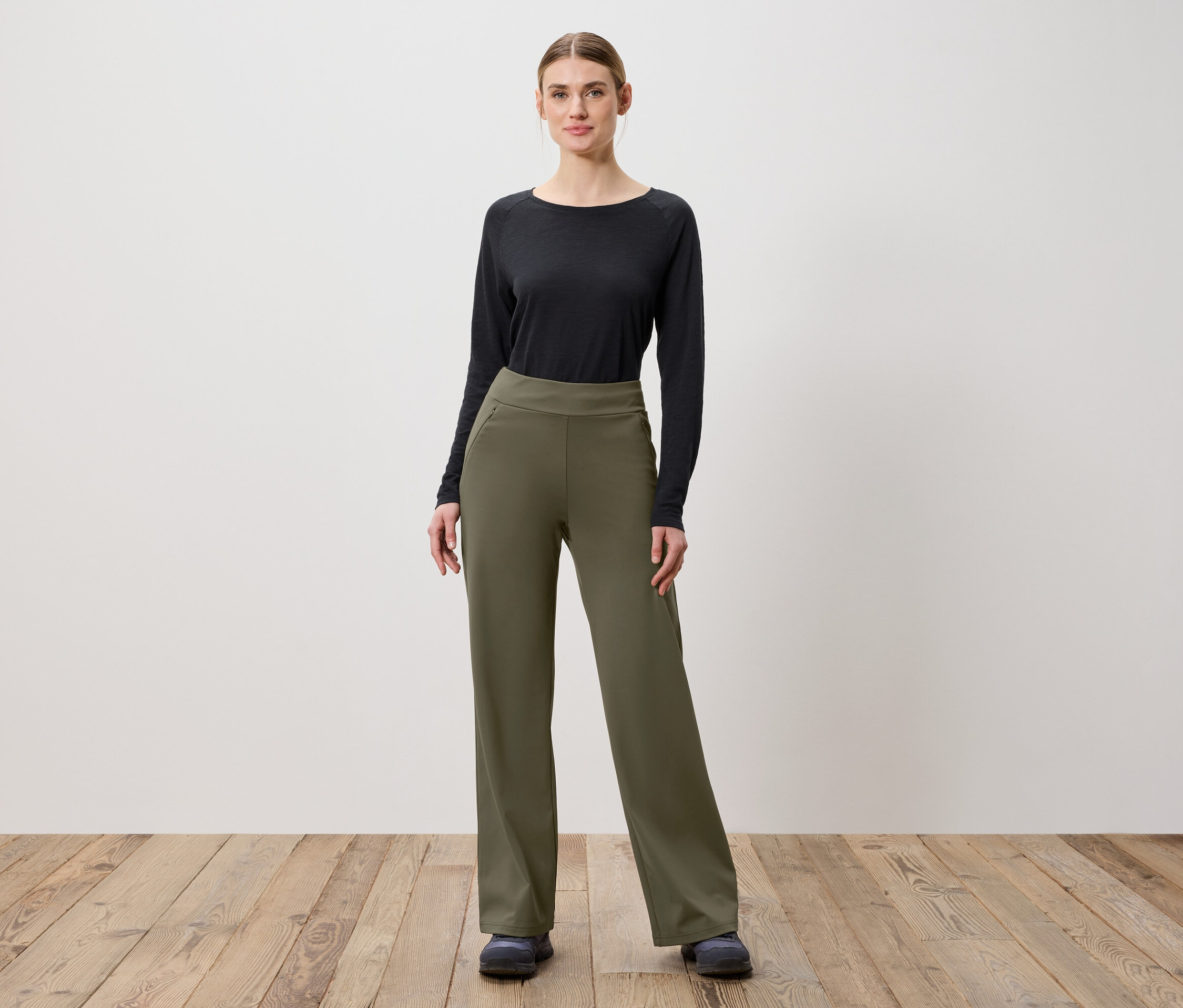 Une femme pose sur un plancher en bois, portant un pantalon vert olive et un haut noir à manches longues.