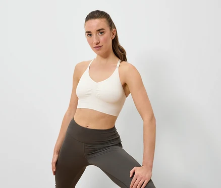 Une femme pose en soutien-gorge de sport crème et leggings gris sur un fond blanc.