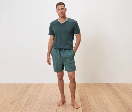 Un homme se tient pieds nus sur un plancher en bois, portant un pyjama vert à manches courtes et un short.
