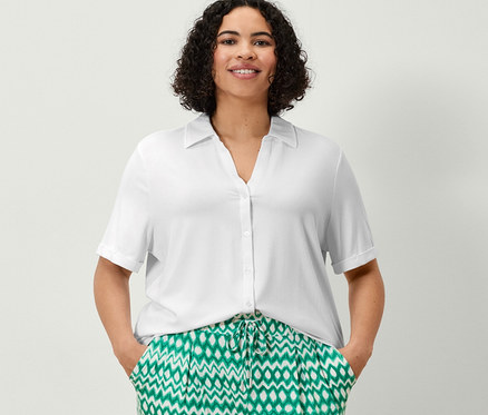 Une femme se tient debout, les mains dans les poches d'un pantalon 3/4 en jersey imprimé et d'un chemisier blanc avec patte de boutonnage.