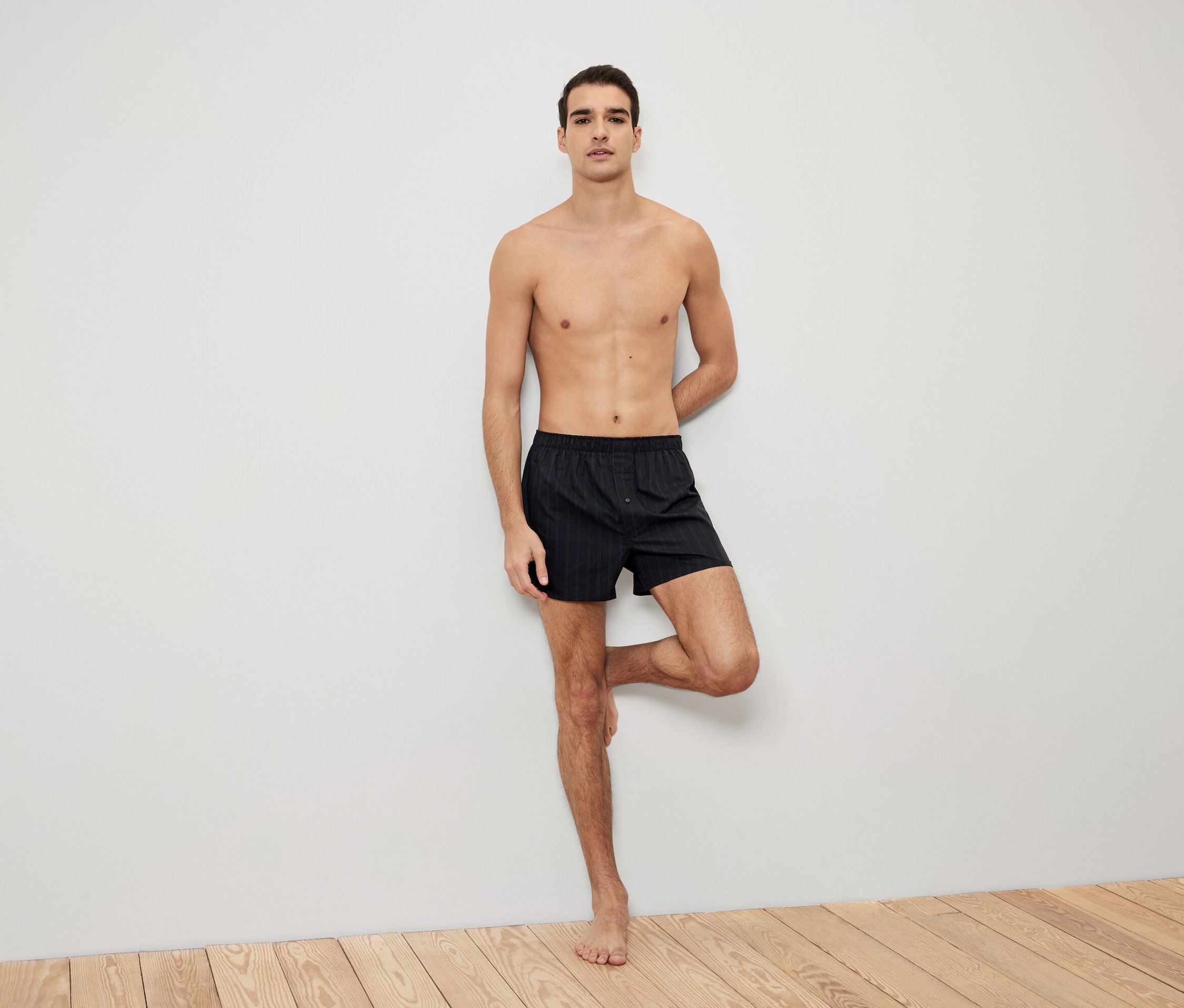 Un homme pose, appuyé contre un mur, en boxeur noir rayé.