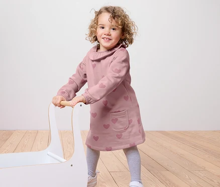 Une petite fille souriante pousse un chariot à jouets blanc. Elle porte une robe rose à motifs de cœurs, des leggings gris et des baskets blanches.