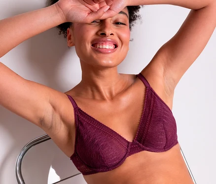 Femme souriante portant un soutien-gorge à armatures en dentelle bordeaux « Leila » Passionata et une culotte assortie, adossée les bras levés.