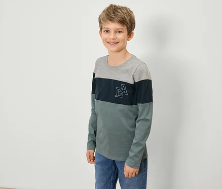 Un garçon portant un t-shirt à manches longues avec un haut gris, une bande bleu marine avec un logo LA et un bas vert, se tient devant un mur blanc.