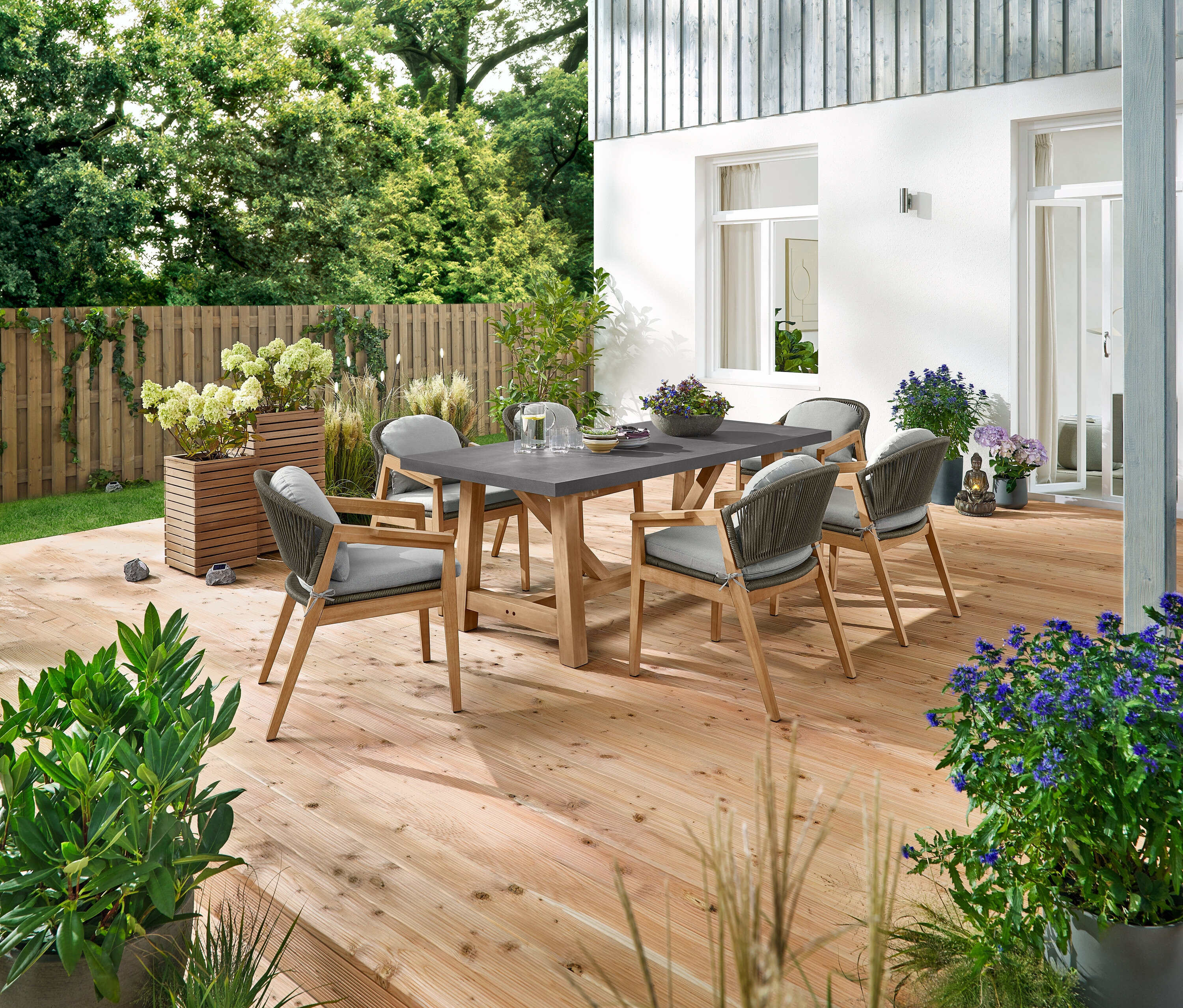 Une table à manger avec un plateau en polyciment et six chaises de jardin confortables avec un tissage textile sont placées sur une terrasse en bois. Les éléments décoratifs sont 3 pierres à LED solaires Premium, 2 colonnes à plantes « Tinus », et 5 piquets à LED solaires.