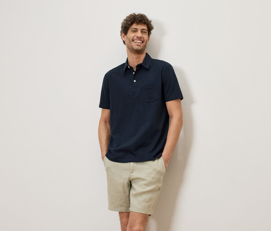 Un homme souriant aux cheveux bouclés se tient devant un mur, portant un polo bleu marine et un short.