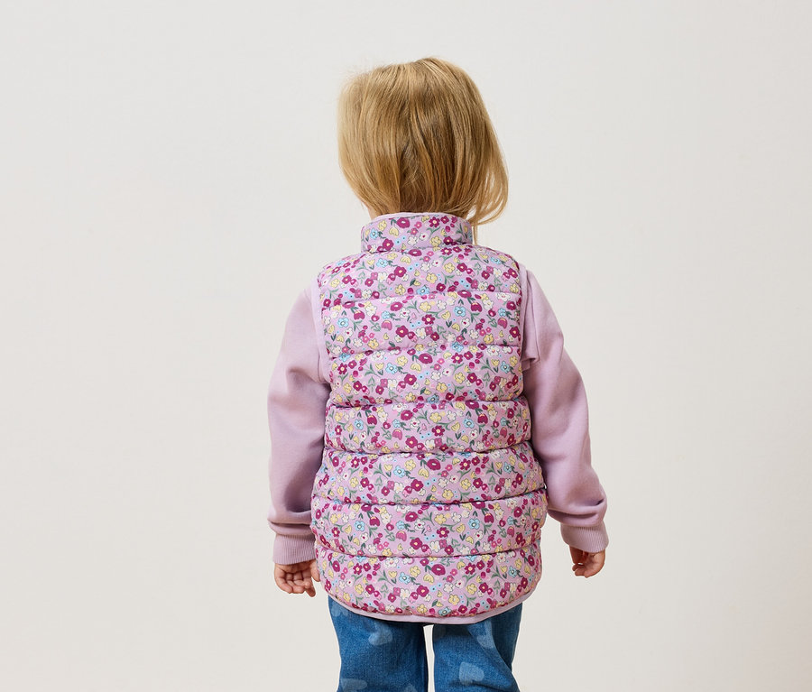 Vue de dos d'un jeune enfant aux cheveux blonds portant un gilet matelassé à fleurs sur un pull rose et un jean.