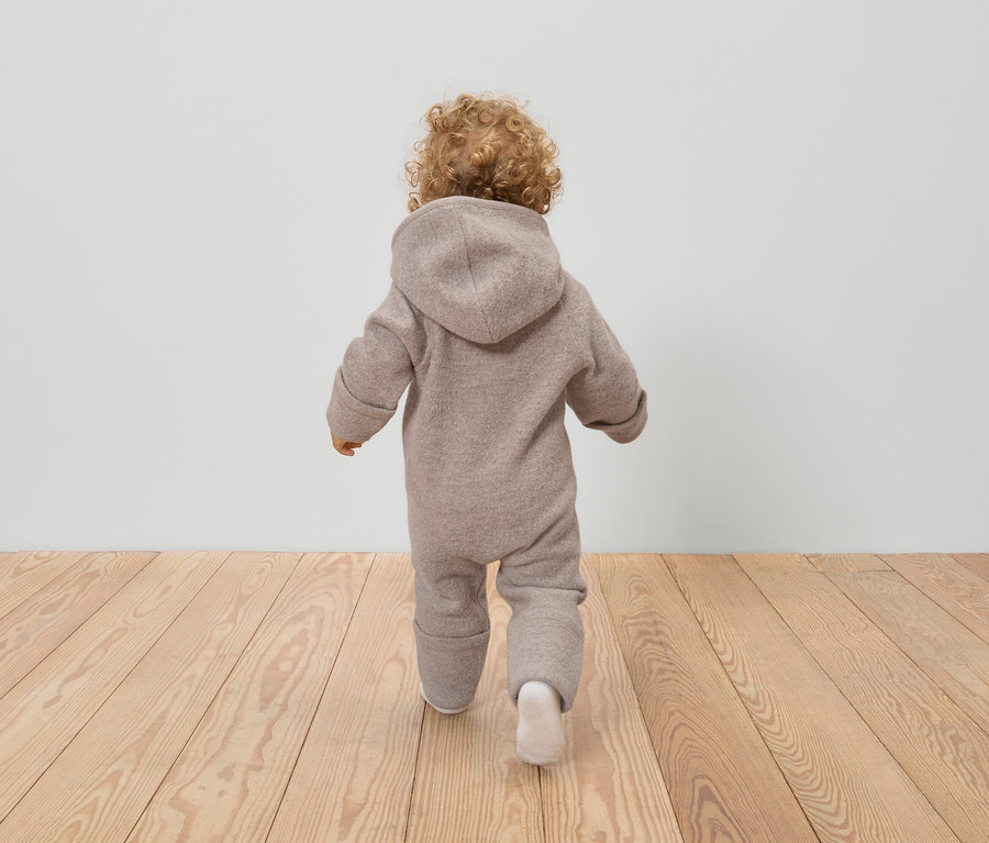 Vue arrière d'un jeune enfant aux cheveux bouclés, vêtu d'une combinaison en laine beige à capuche et de chaussettes blanches, debout sur un plancher en bois.