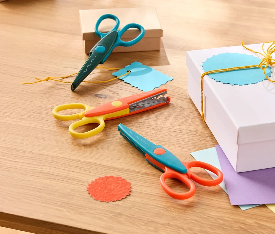Trois paires de ciseaux décoratifs bleu-vert, jaune-orange et bleu-orange sont posées sur une table en bois à côté de boîtes cadeaux et de papier coloré.